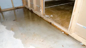 hoa-and-condo-water-damage-who-is-responsible-in-maryland_11zon - First & Last Restoration Wet concrete floor in a partially framed room under construction, with standing water near an open doorway and exposed studs along the walls.