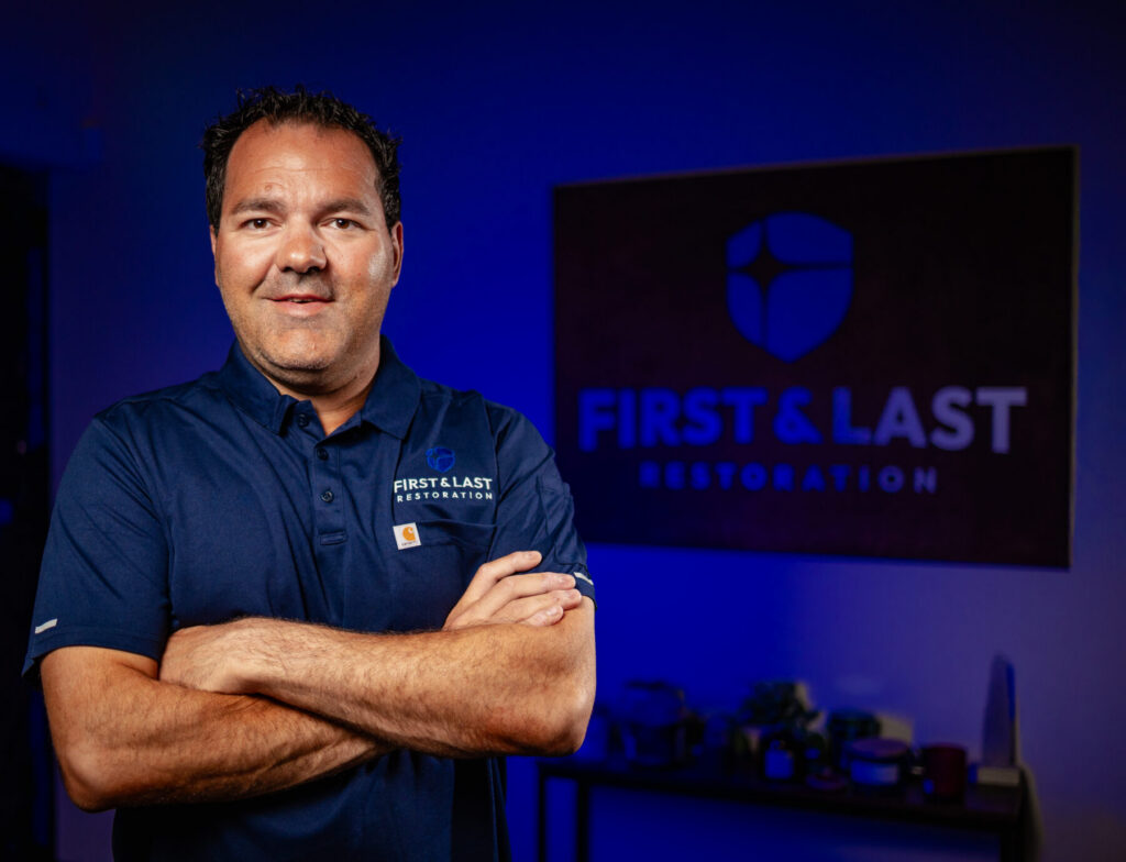 Professional man with arms crossed in a blue-lit room, wearing a First & Last Restoration polo shirt, standing in front of a sign displaying the company logo.